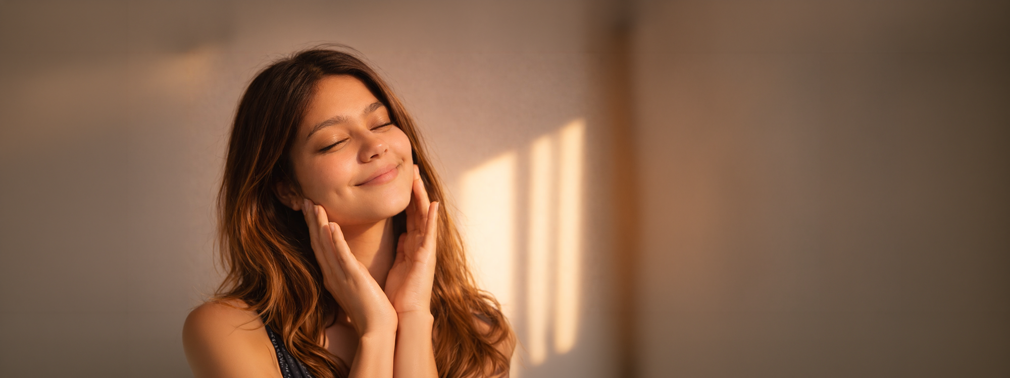 Woman with long brown hair standing in a softly lit room, hands on face.